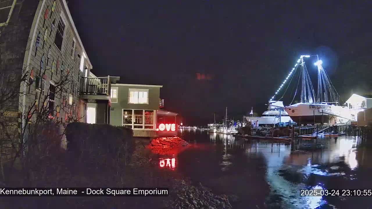 A nighttime waterfront scene shows a building decorated with nautical flags, a large sailboat docked, and illuminated “LOVE” letters near the water’s edge.