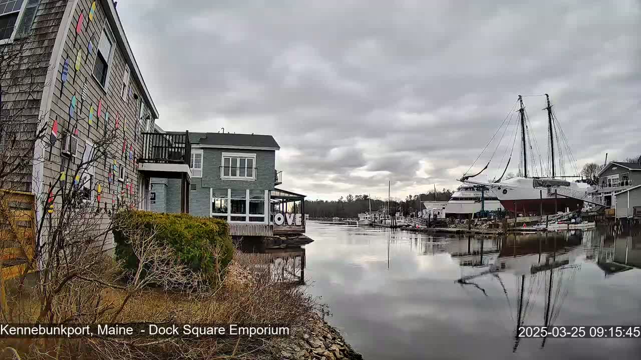 A calm river reflects a large sailboat and other boats under an overcast sky, with buildings and vegetation lining the riverbank.