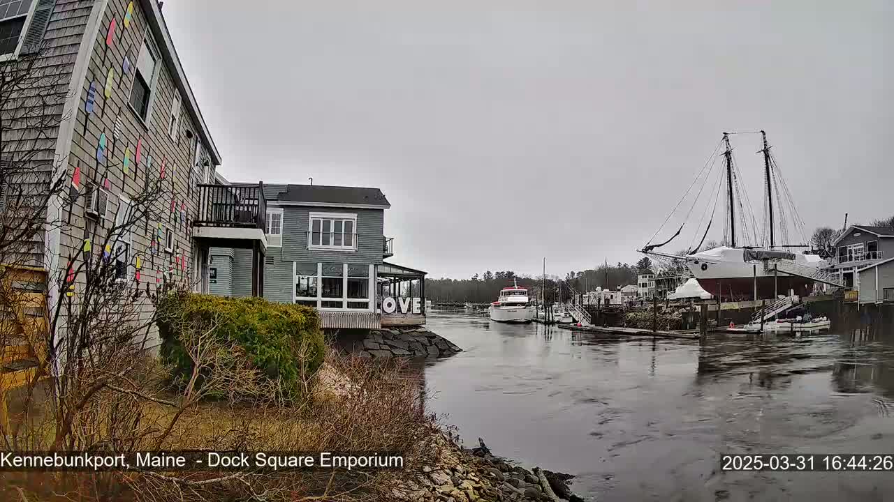 A gray day shows a waterfront scene with a building decorated with colorful nautical flags, another building with a "LOVE" sign, several boats including a large sailing ship, and calm water.