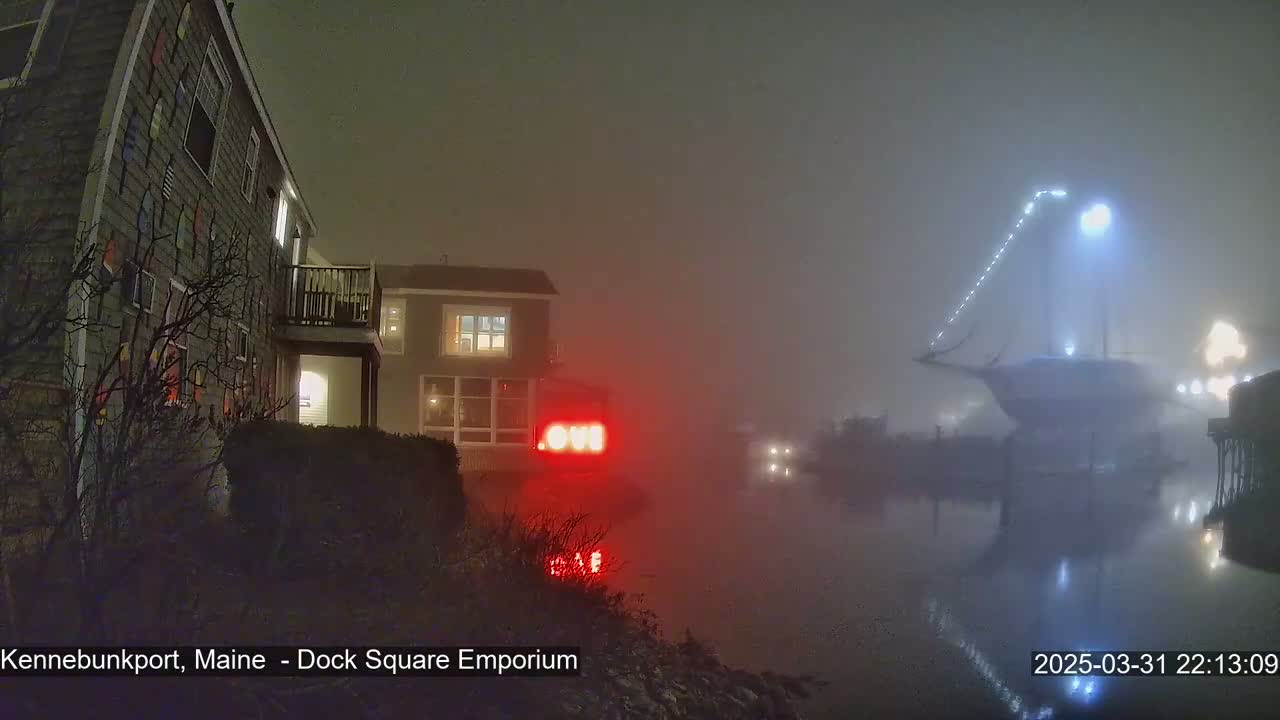 A foggy night scene shows a waterfront with buildings, one adorned with colorful nautical decorations, and a large boat illuminated by lights in the water.