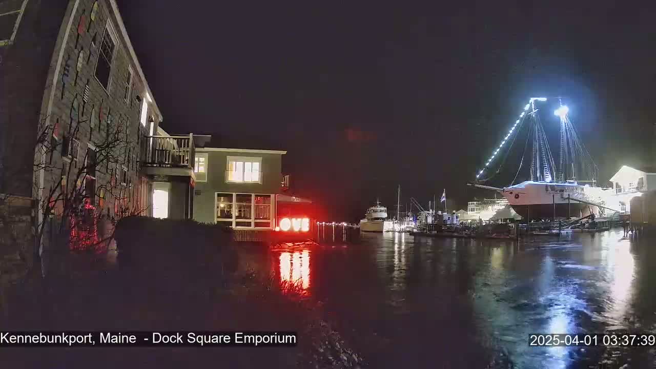 A nighttime scene shows a building with a decorative exterior, illuminated by lights, near a harbor where boats are docked, under dark, calm conditions.