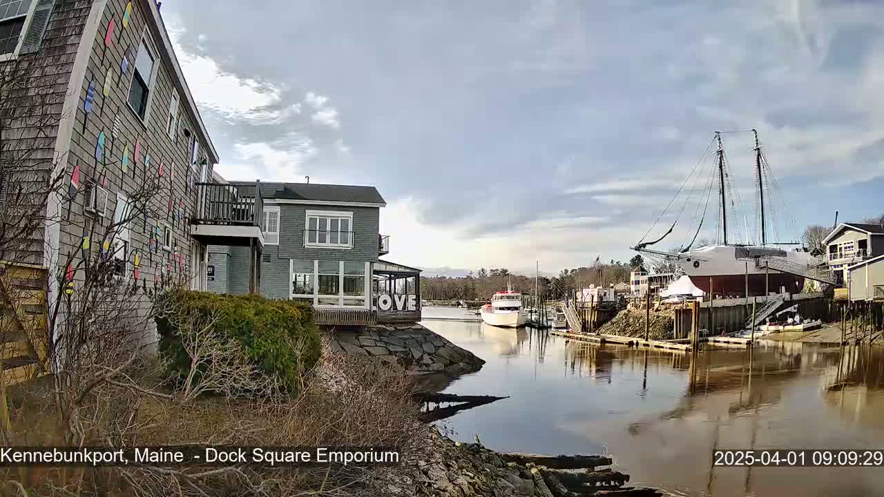 A partly cloudy day reveals a waterfront scene with colorful buildings, a large sailboat docked, and a smaller motorboat on calm water.