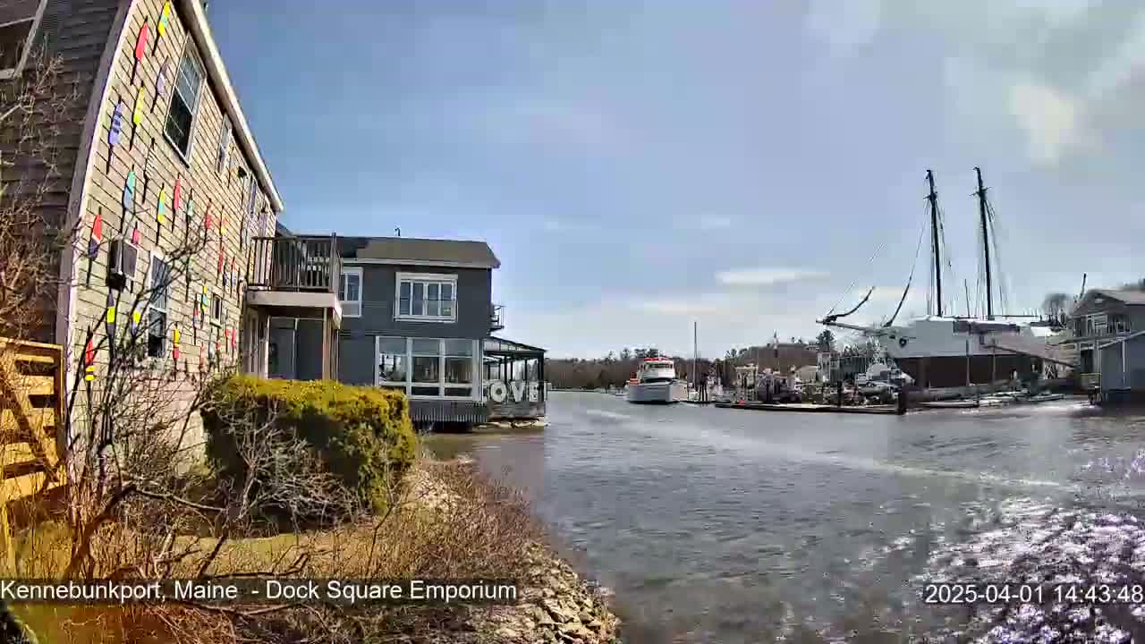A waterfront scene on a sunny day shows a building with colorful decorations, another building with large windows, a small motorboat passing by, and a large sailboat docked nearby.