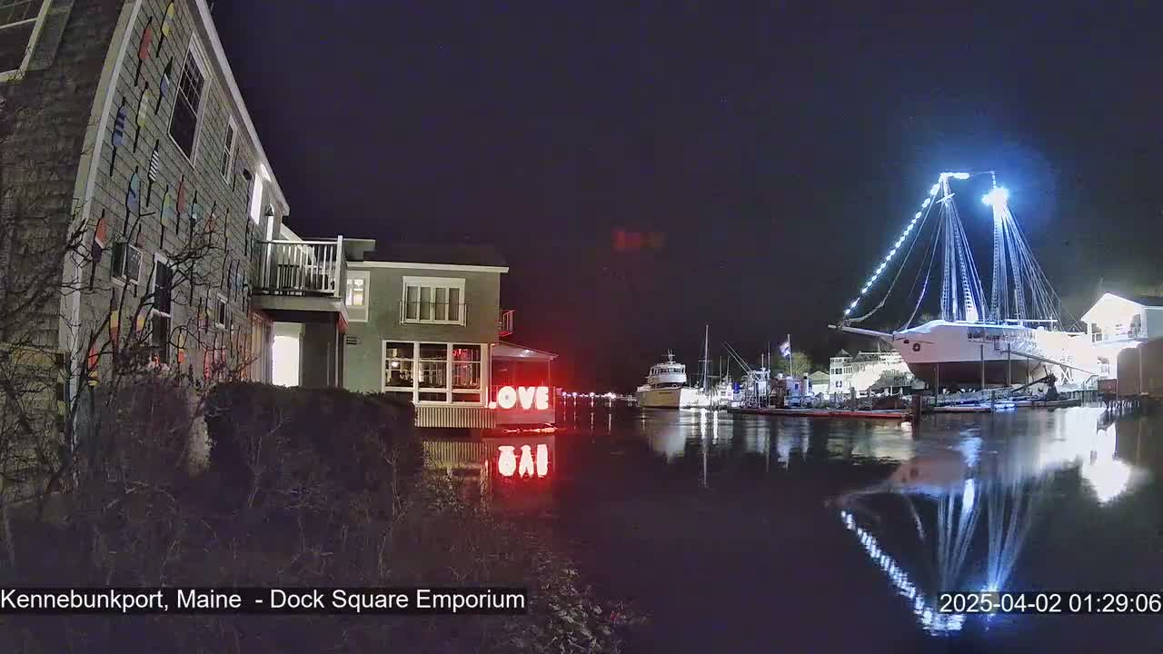 A nighttime view of a waterfront scene showing a building decorated with nautical flags, a large illuminated ship docked, and the word "LOVE" in lights reflected in calm water.
