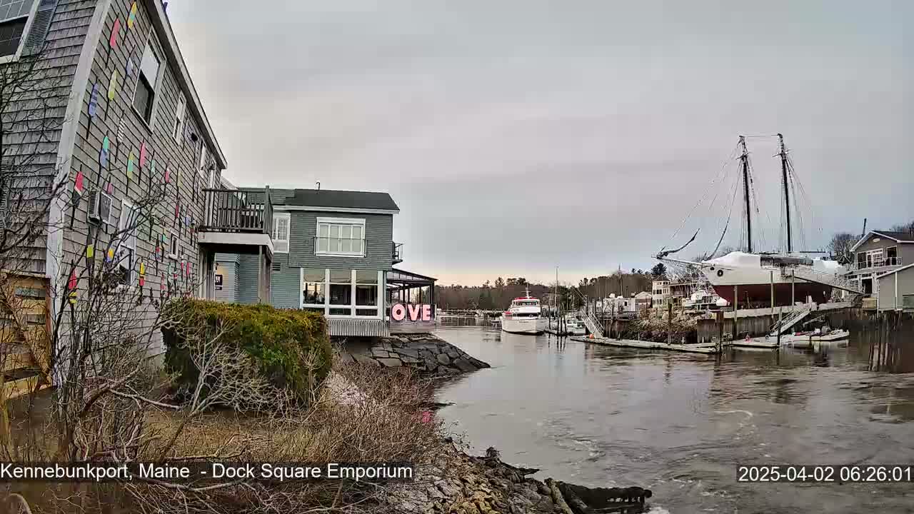 A waterfront scene on an overcast day shows a building decorated with colorful nautical flags, another building with a "LOVE" sign, a docked sailboat, and a motorboat on the water.