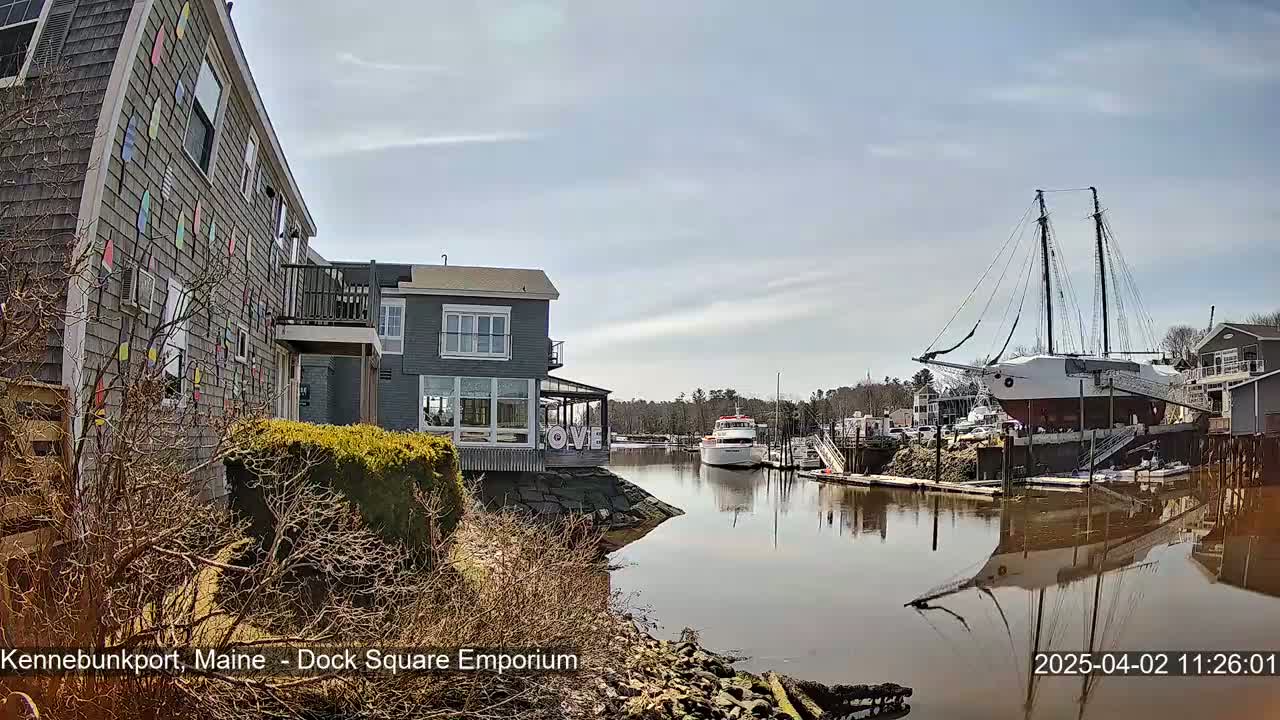 A calm waterway reflects buildings and a large sailboat under a mostly sunny sky.