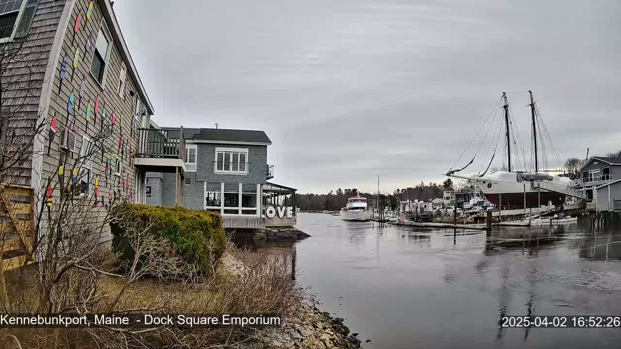 A waterfront view on an overcast day shows a gray building with colorful nautical flags on its side, another gray building across a calm waterway, a large sailing ship docked nearby, and a smaller passenger boat further down the waterway.