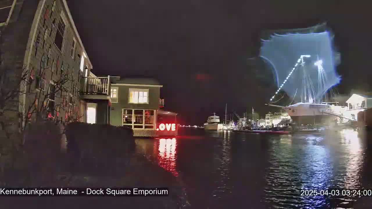 A nighttime waterfront scene shows a building with illuminated red "LOVE" letters, a sailboat with a light display above it, and other boats moored in the water.