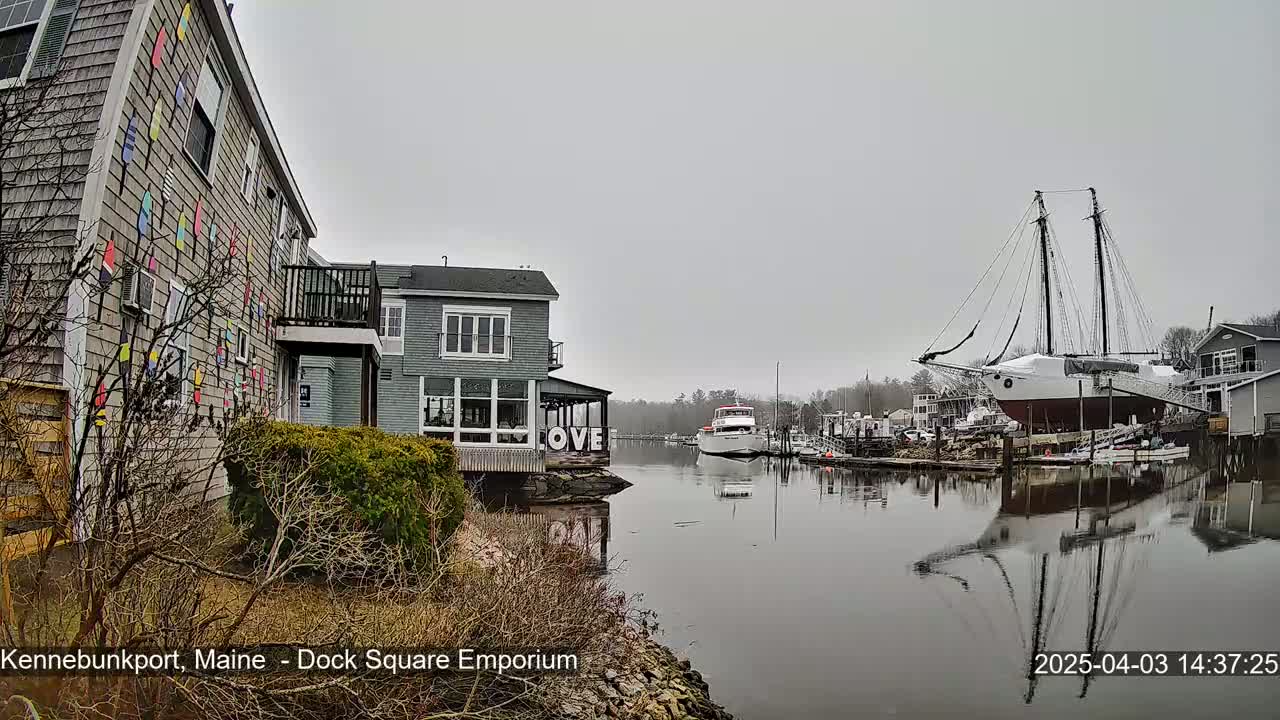 A calm, overcast day reveals a waterfront scene with buildings, a large sailing ship docked, smaller boats, and calm water reflecting the surroundings.