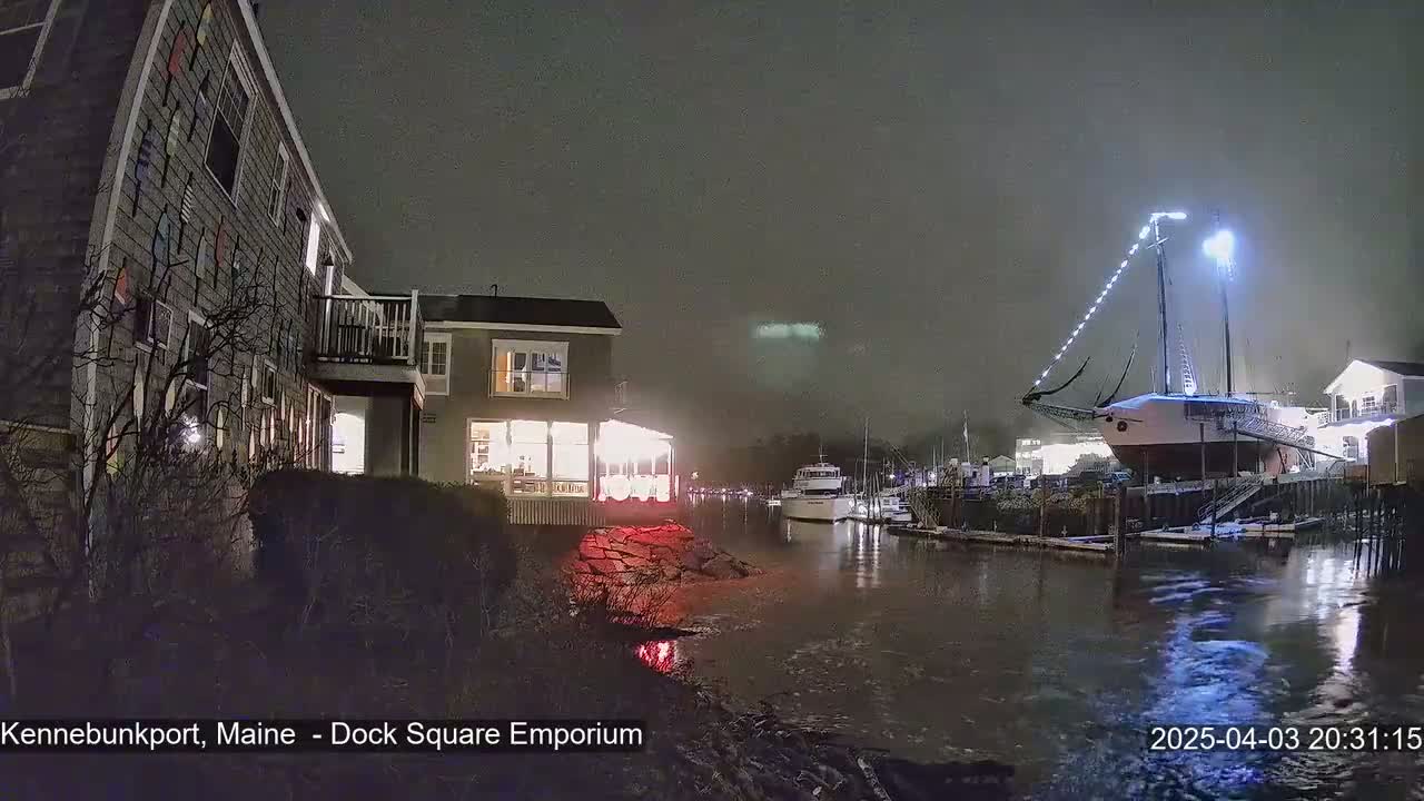 A nighttime scene shows a snow-dusted sailboat docked near lit buildings and other boats in calm water.