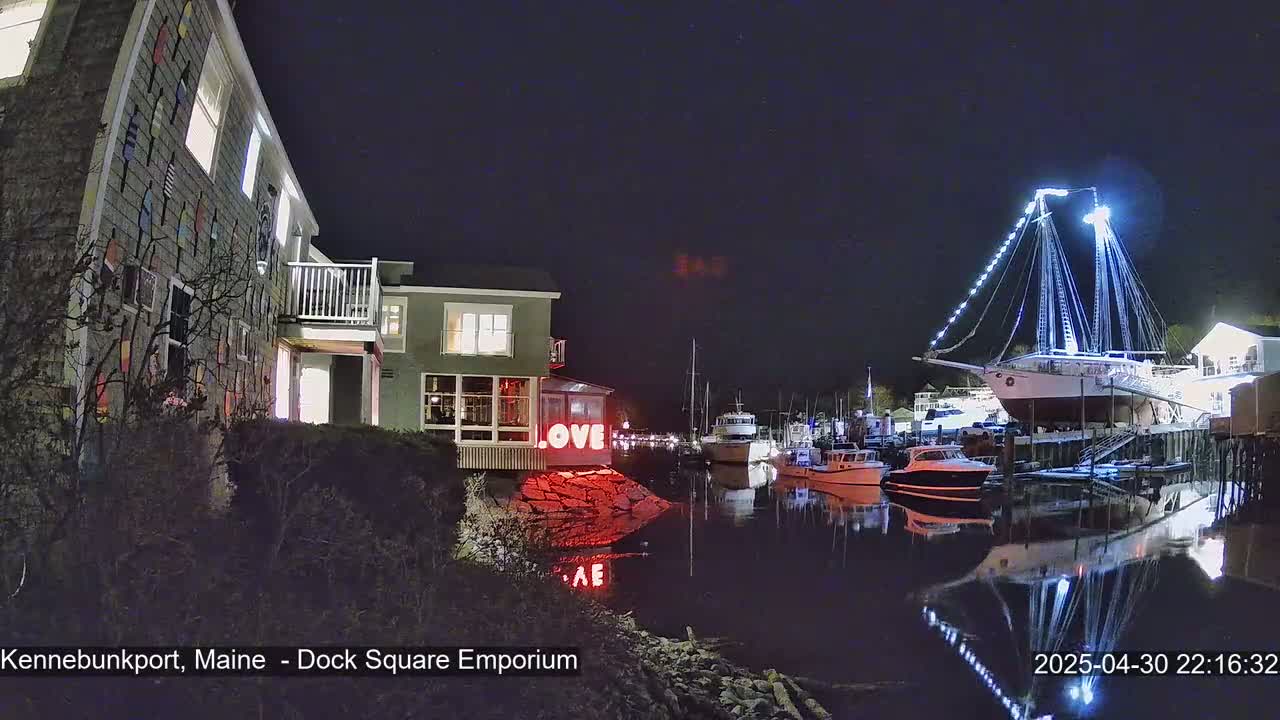 At night, a waterfront scene shows a building decorated with nautical flags, a lit-up sailboat, and several smaller boats moored in calm water, reflecting the lights.