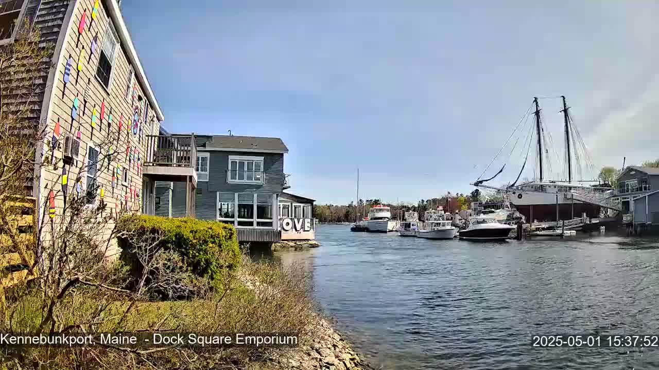 A waterfront scene on a sunny day shows a building decorated with colorful nautical flags, other buildings, and several boats including a large sailing ship moored at a dock.