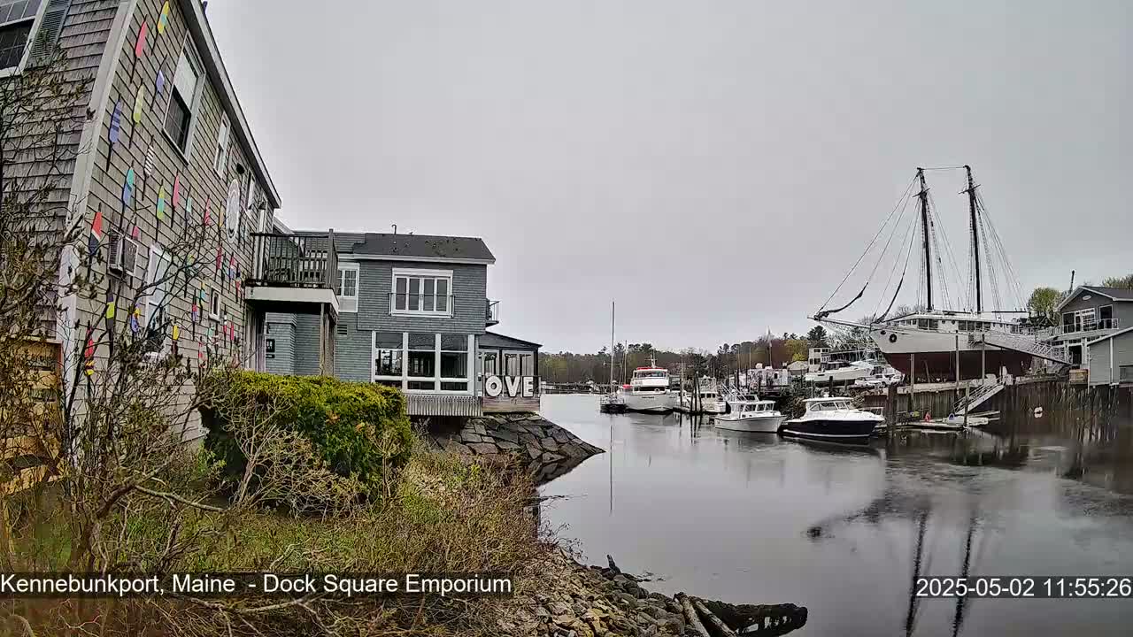 A harbor scene on an overcast day shows a gray building with colorful pennants on its side, other buildings, several boats including a large sailboat on a cradle, and calm water reflecting the sky.