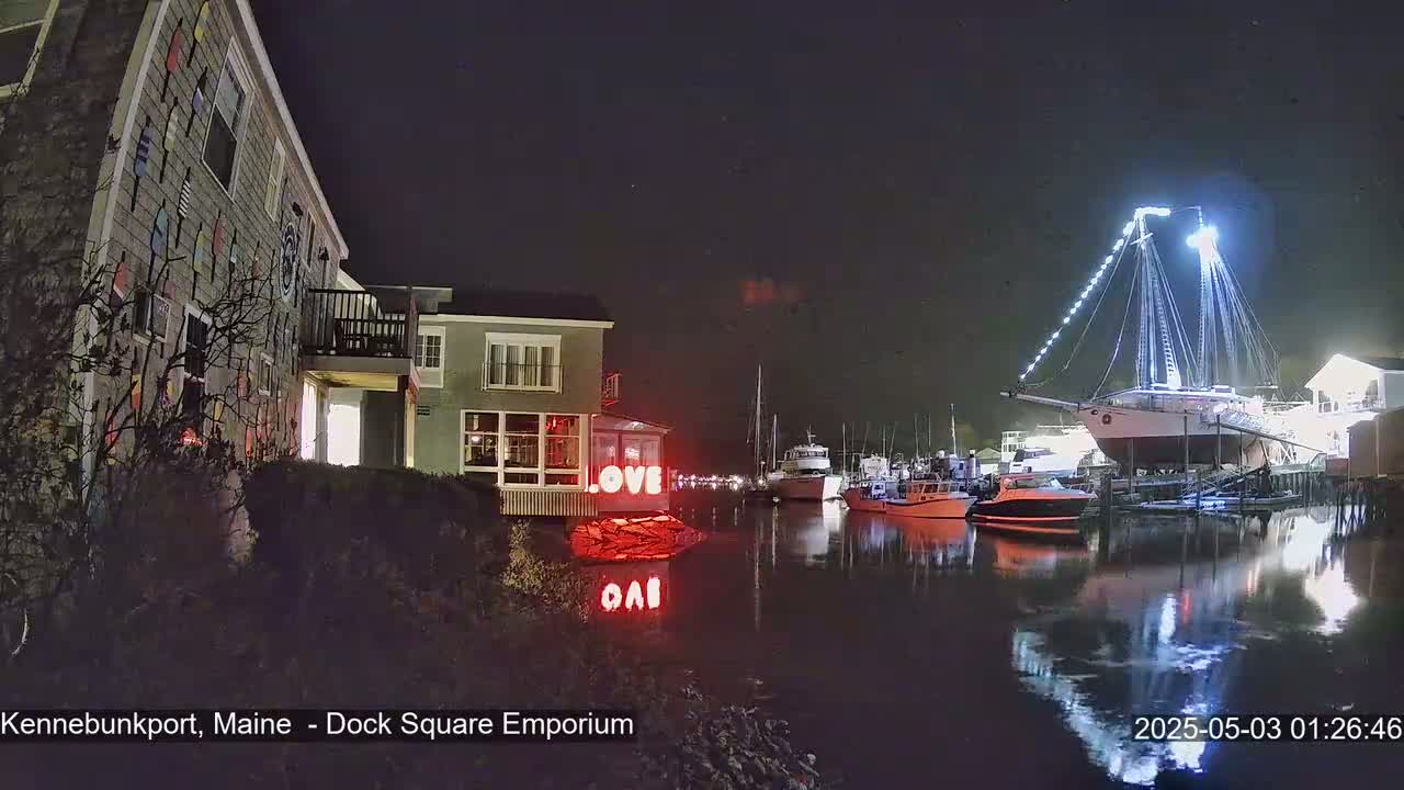 At night, a brightly lit sailboat is docked in a harbor next to buildings, one of which displays illuminated "LOVE" signage.