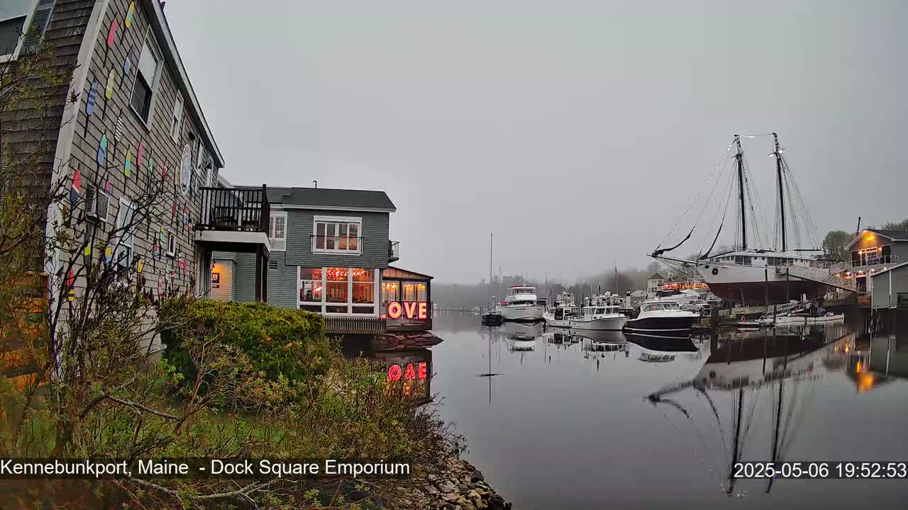A waterfront scene on an overcast day shows a building with colorful decorations next to a building with illuminated windows displaying the word "LOVE," several boats moored at the water's edge, and a large sailing ship.