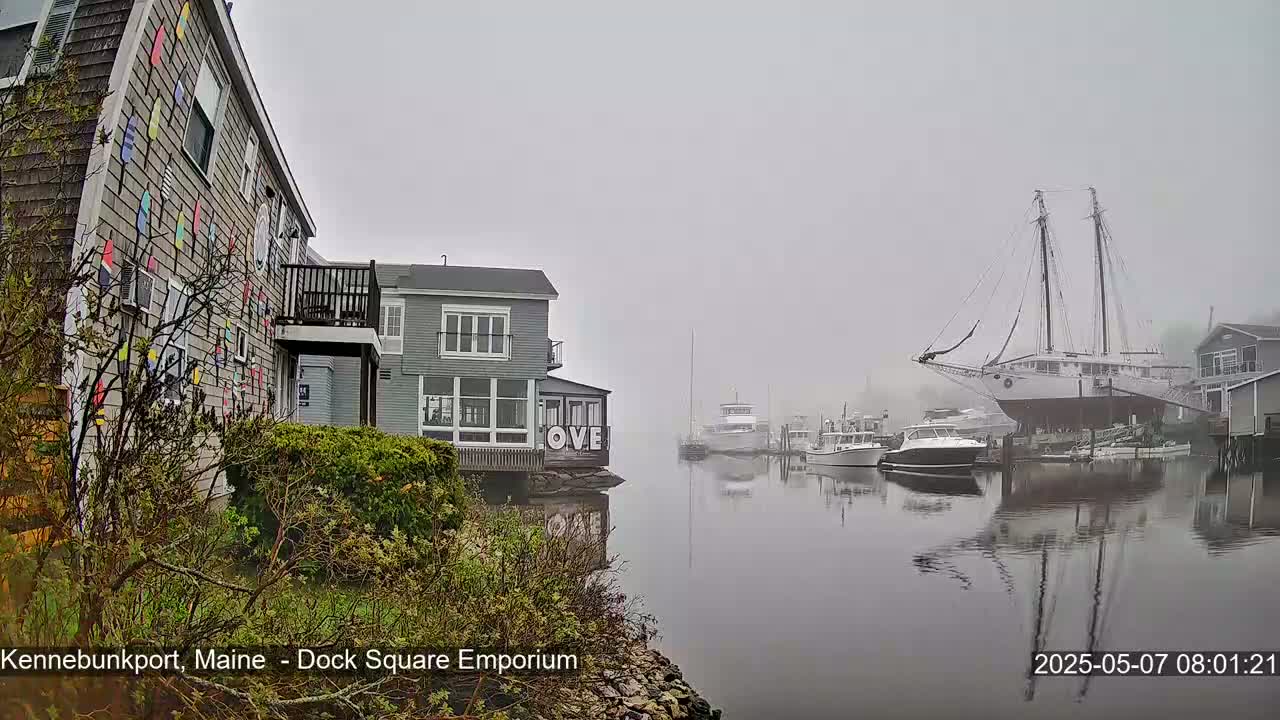 A foggy waterfront scene shows a building with colorful paddle-shaped decorations, another building with a porch, several boats, and a large sailboat on land.