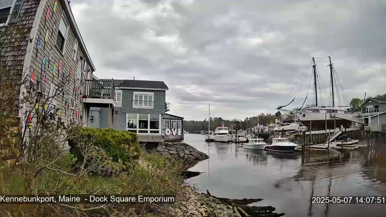 A harbor scene under an overcast sky shows a building decorated with colorful pennants, other buildings, several boats, and a large sailboat on a dry dock.