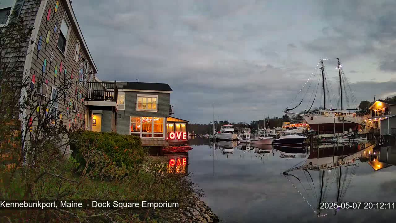 A calm harbor at dusk shows a building with colorful nautical flags on its exterior, a restaurant with illuminated "LOVE" signage, several boats, and a large sailing ship reflected in the water.