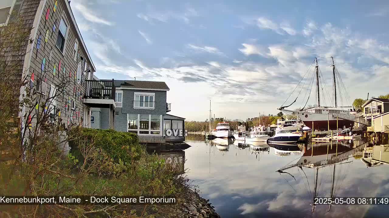 A calm waterway reflects a partly cloudy sky, several boats, and buildings, including one with colorful decorations on its side.