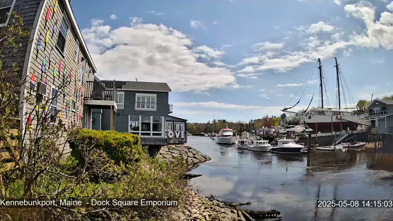A waterfront scene on a partly sunny day shows a gray building with colorful decorations, other buildings, and several boats docked along a calm waterway.