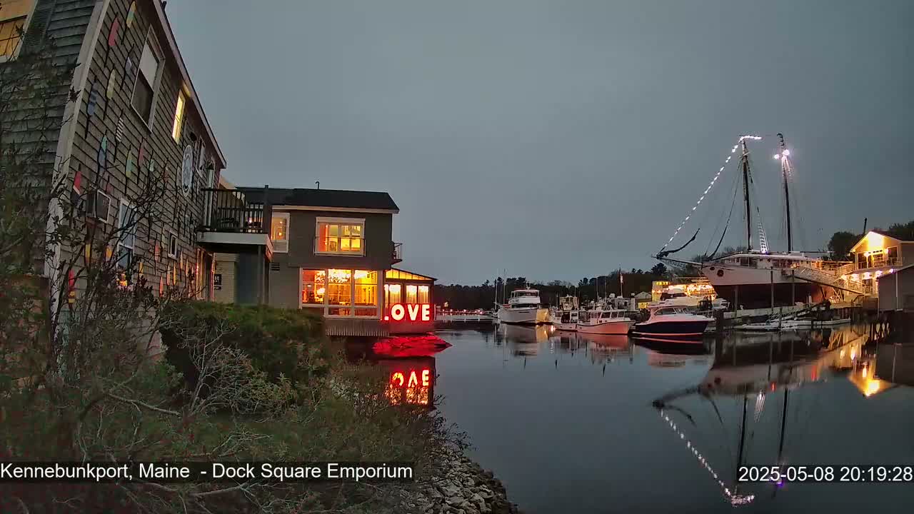 Under a twilight sky, illuminated buildings and boats line a calm harbor, with a large sailboat prominently featured.