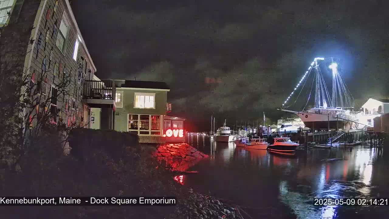 A nighttime harbor scene shows a large sailboat illuminated with lights, other boats in the water, buildings on the shore, and a red "LOVE" sign near a building.