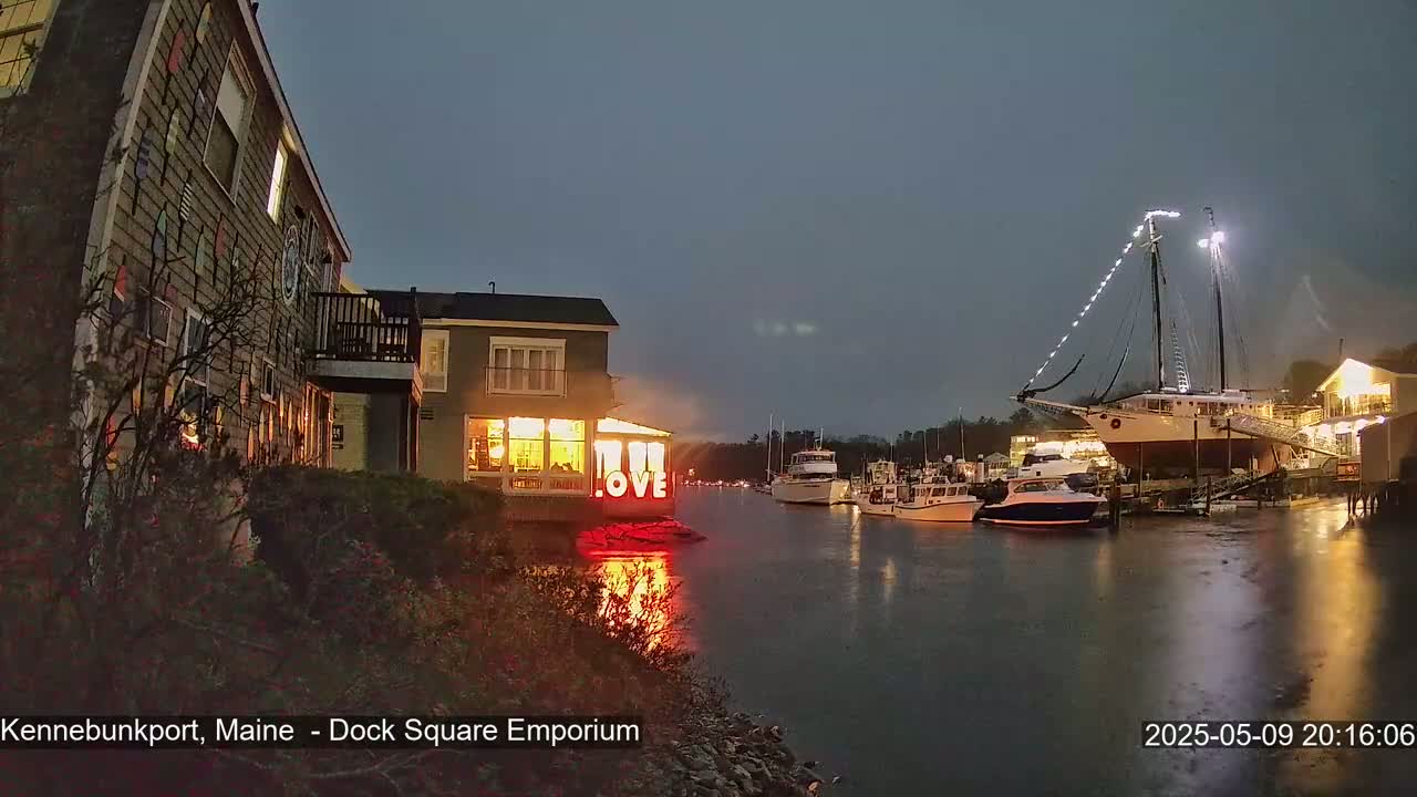 A nighttime waterfront scene shows a building with illuminated "LOVE" signage, several boats moored at a dock, and rain falling under a dark sky.