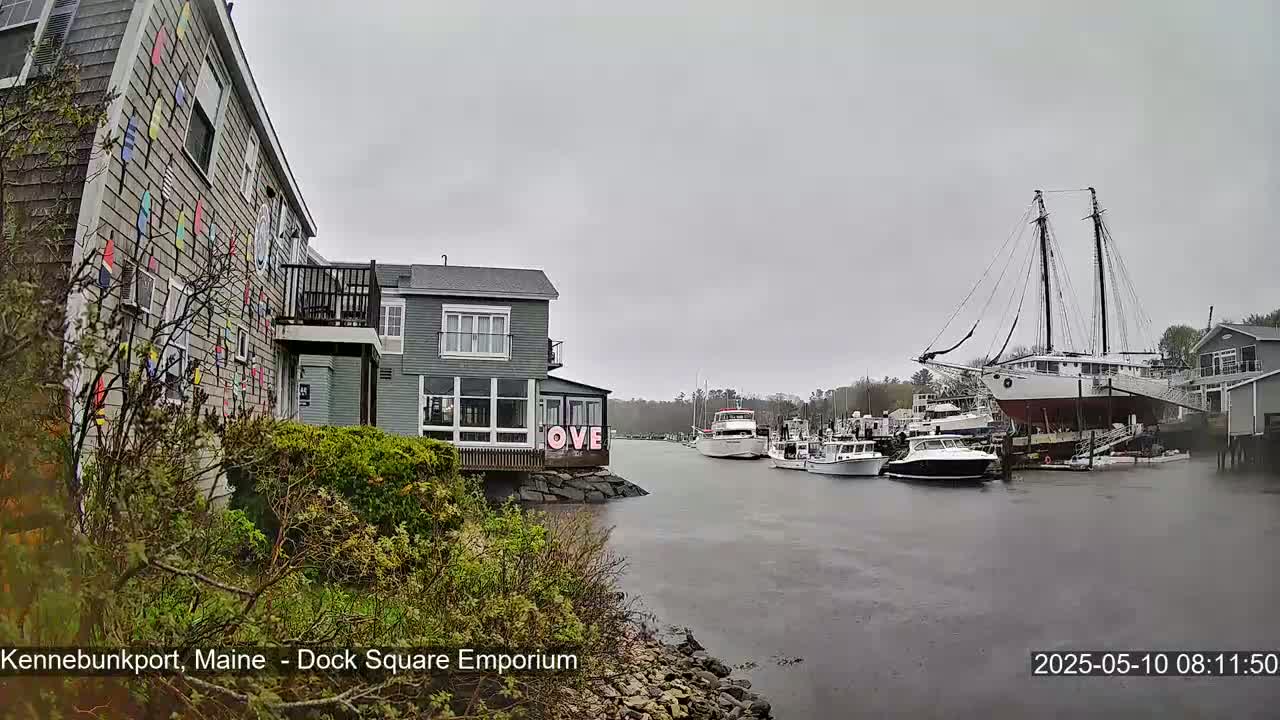 A gray, overcast day shows a waterfront view with several boats, including a large sailing ship, moored in a calm waterway, next to buildings, one of which has colorful nautical flags on its side.