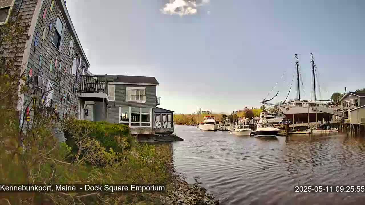 A waterfront view on a partly sunny day shows a gray building with colorful accents, other buildings, and several boats in a calm waterway.