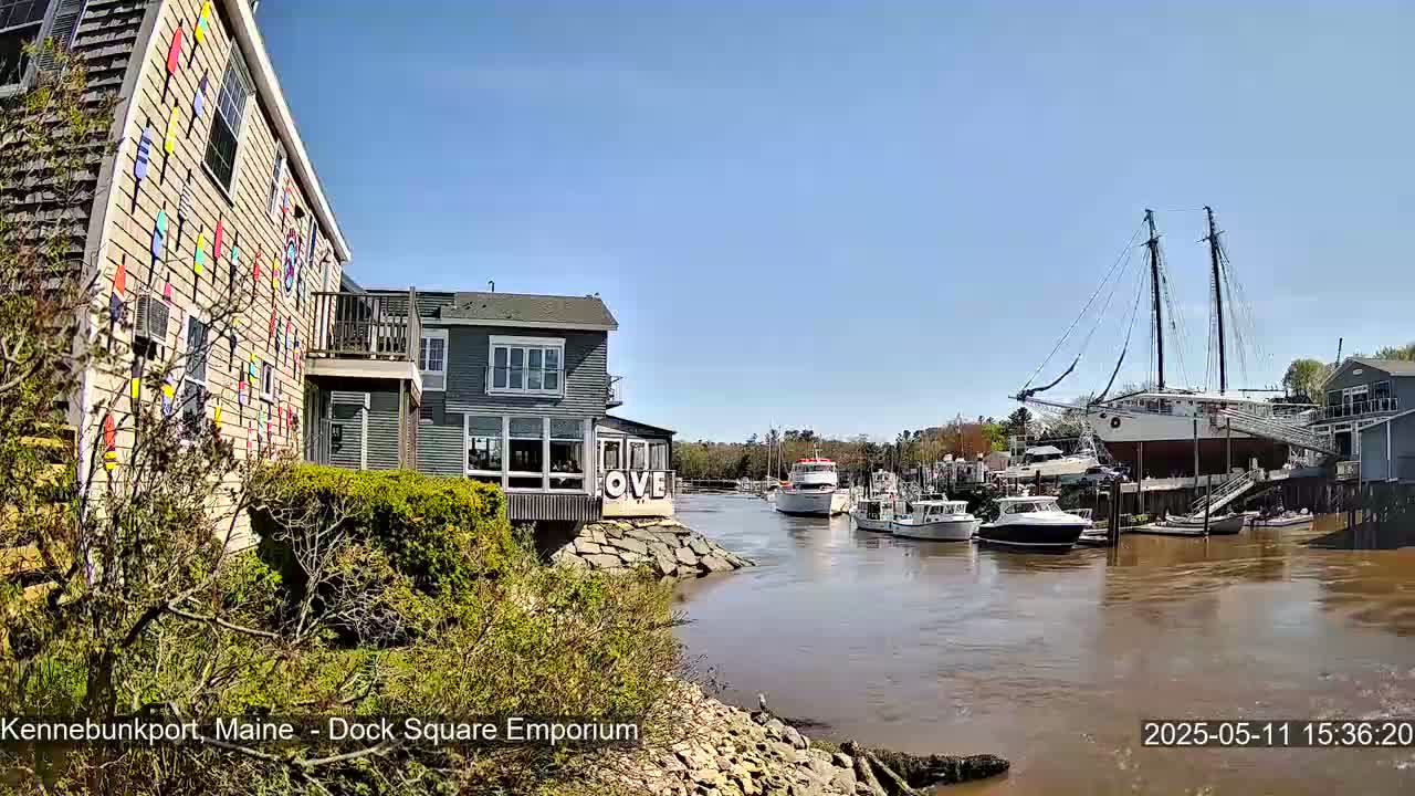 A waterfront scene on a sunny day shows a harbor with several boats, including a large sailing ship, and buildings with colorful accents.