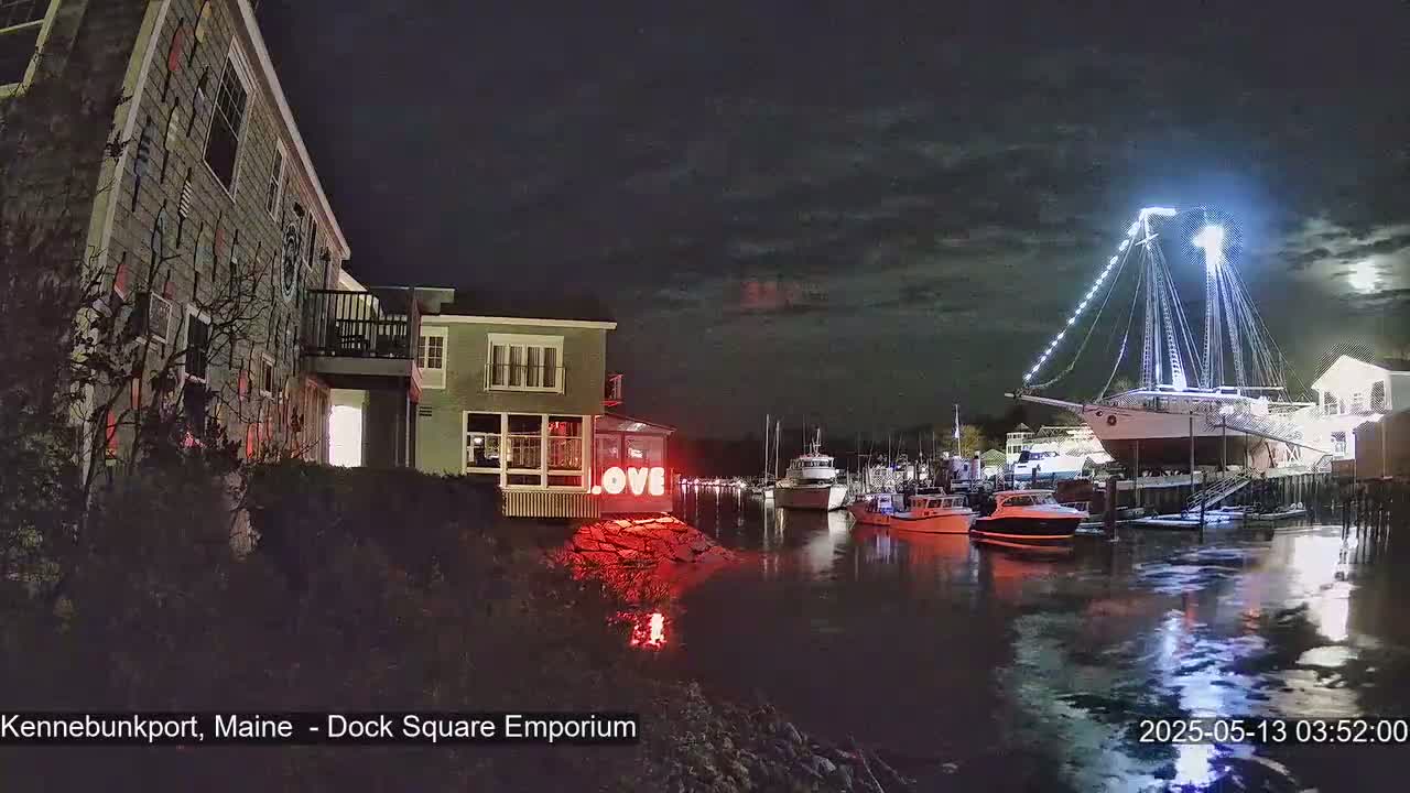 A nighttime harbor scene shows a large illuminated sailboat docked near several smaller boats and buildings, under a partly cloudy sky.