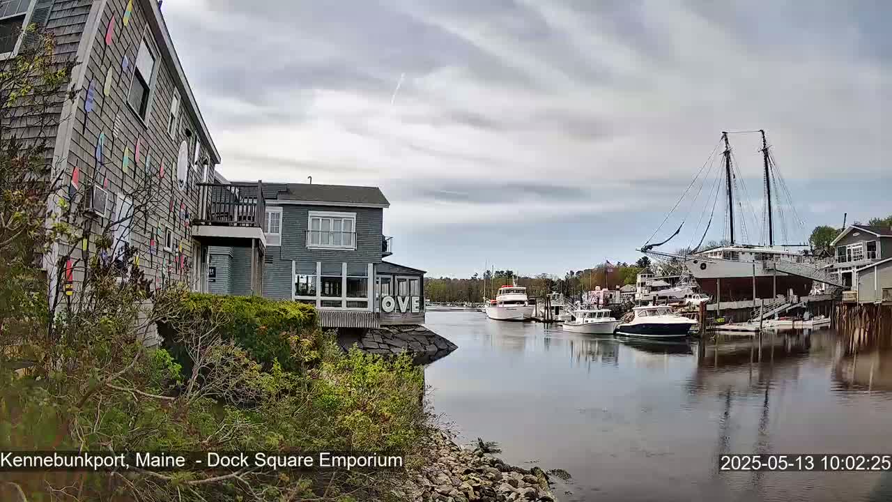 A waterfront scene on a cloudy day shows a gray building with colorful accents, several boats moored in calm water, and other buildings and trees in the background.