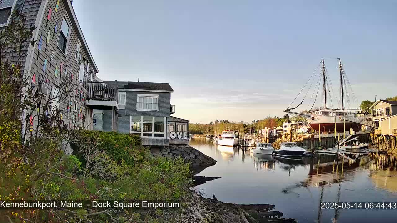 A calm harbor scene at sunrise shows a waterfront building with colorful painted siding, other buildings, several boats, and a large sailboat on a dry dock.