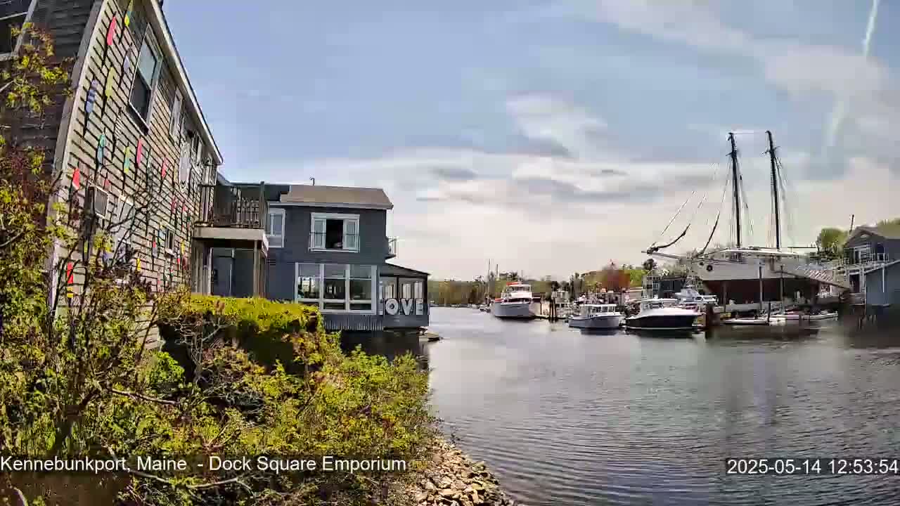 A partly cloudy day reveals a waterfront scene with several boats, including a large sailboat on land, docked along a calm waterway, near buildings with colorful exterior decorations.