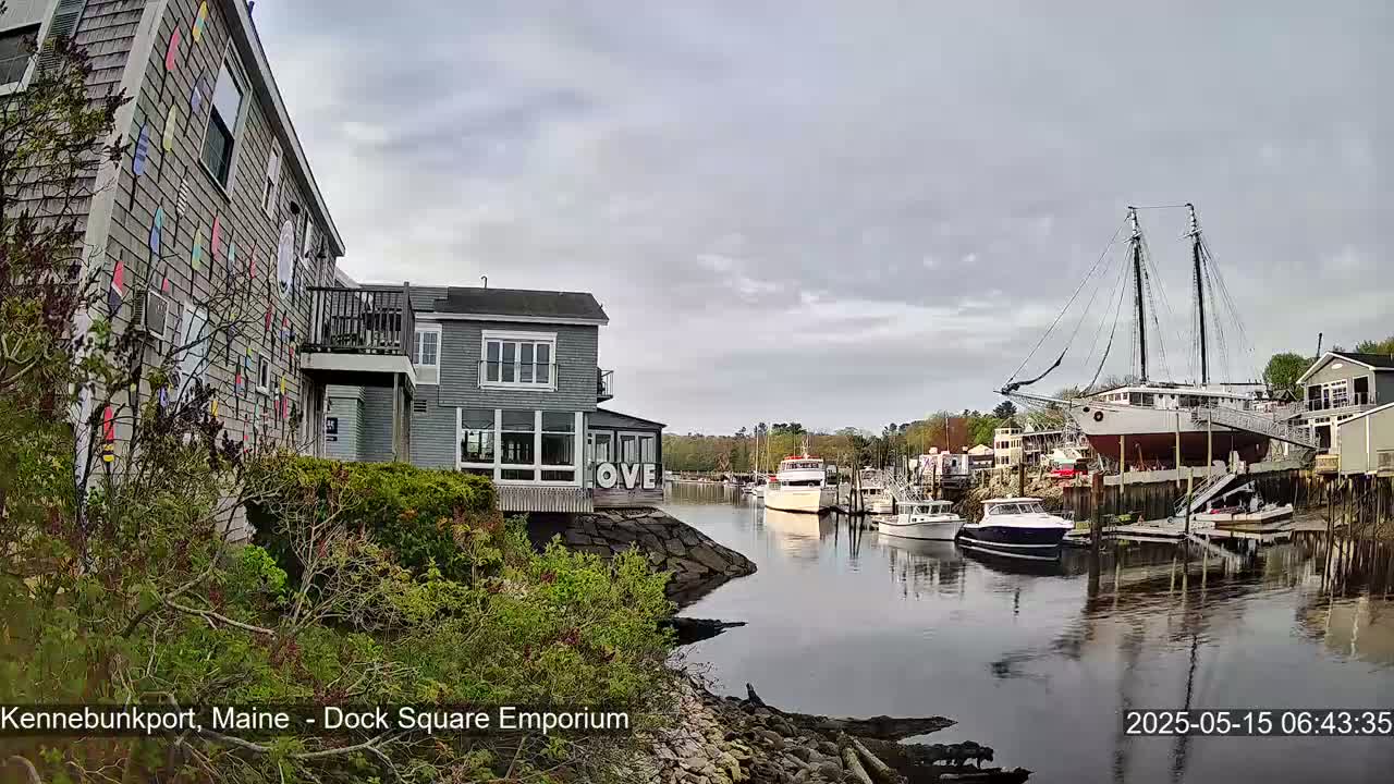 A gray building with colorful nautical flags adorns its side, overlooking a calm harbor with several boats, including a large sailboat on land, under an overcast sky.