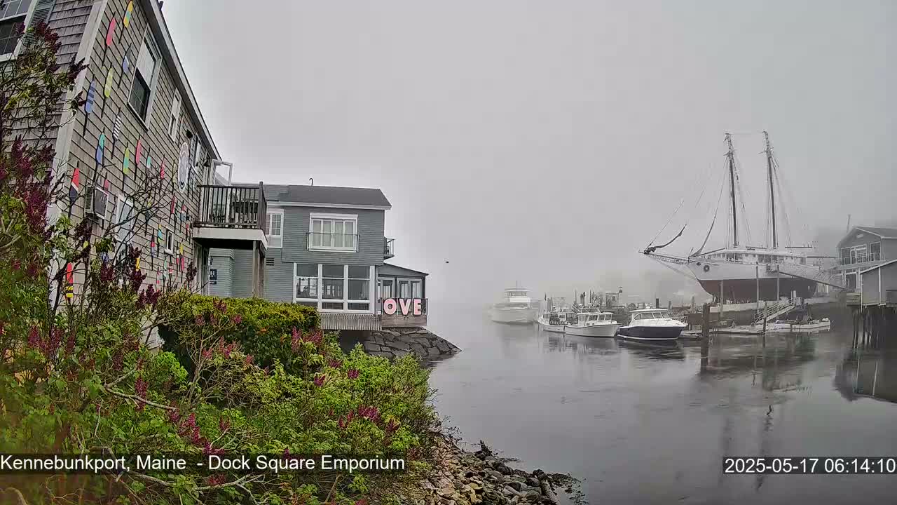 A foggy harbor scene shows a large sailboat on land, several smaller boats in the water, and buildings along the shore, one with colorful decorations on its exterior.