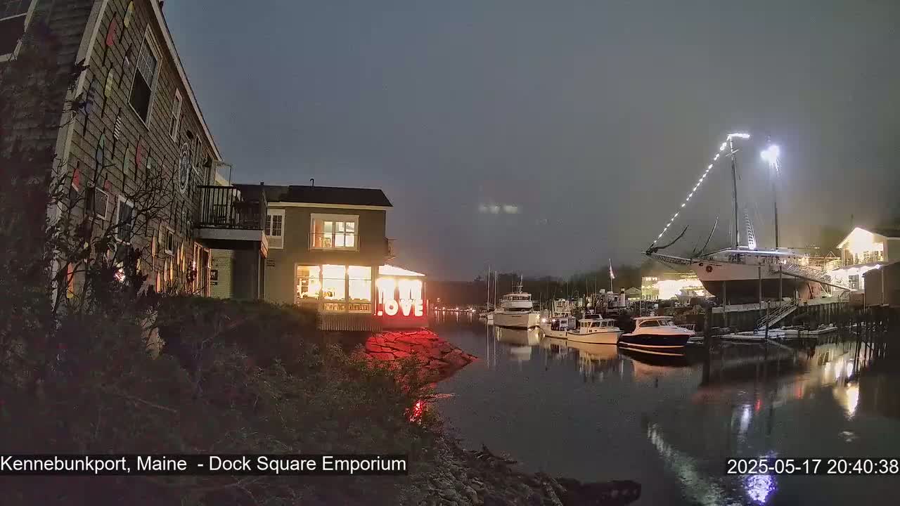 A nighttime harbor scene shows a building illuminated with a "LOVE" sign, several boats docked, and a large sailboat lit up at the pier under an overcast sky.