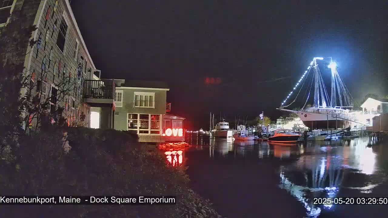 At night, a brightly lit sailboat is docked in a harbor next to buildings, one of which displays illuminated "LOVE" letters.