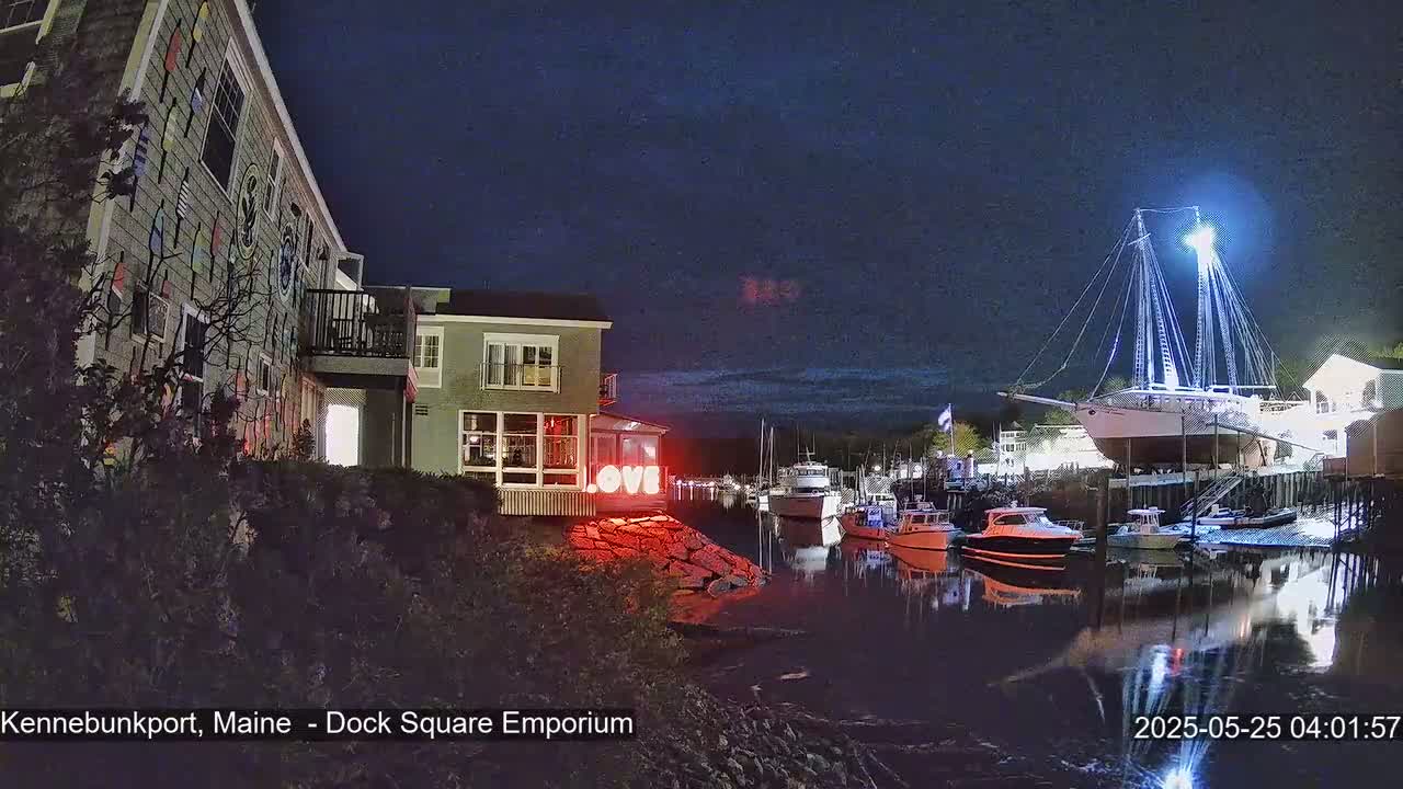 A brightly lit sailboat is moored in a harbor at night, near a building decorated with nautical-themed siding and a lighted “LOVE” sign.