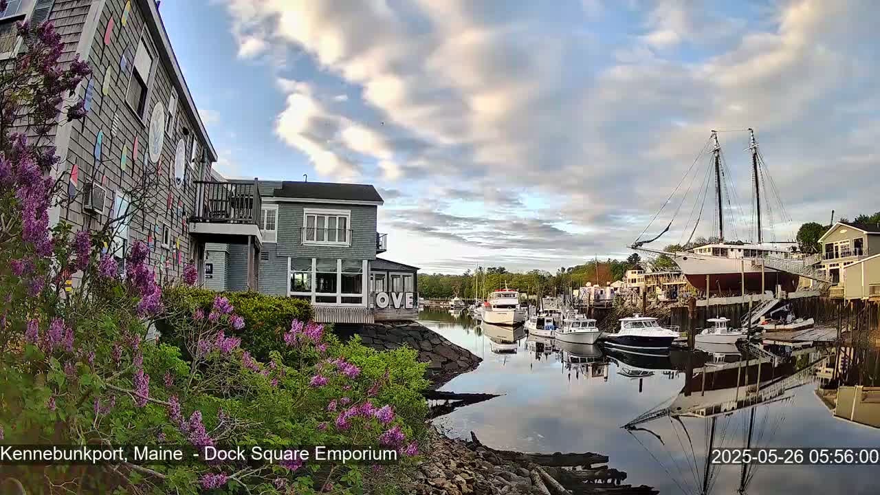 A calm harbor scene with several boats, a large sailing ship, and buildings, all under a partly cloudy sky.