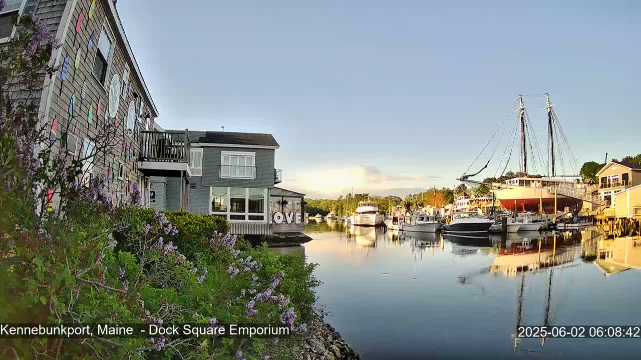 A calm harbor scene at sunrise features a colorful building, several boats, and a large sailing ship docked near a waterfront building.
