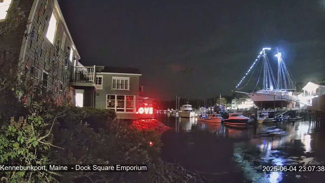 A large illuminated sailboat is docked at a waterfront at night, next to several smaller boats, near a building with a "LOVE" sign.