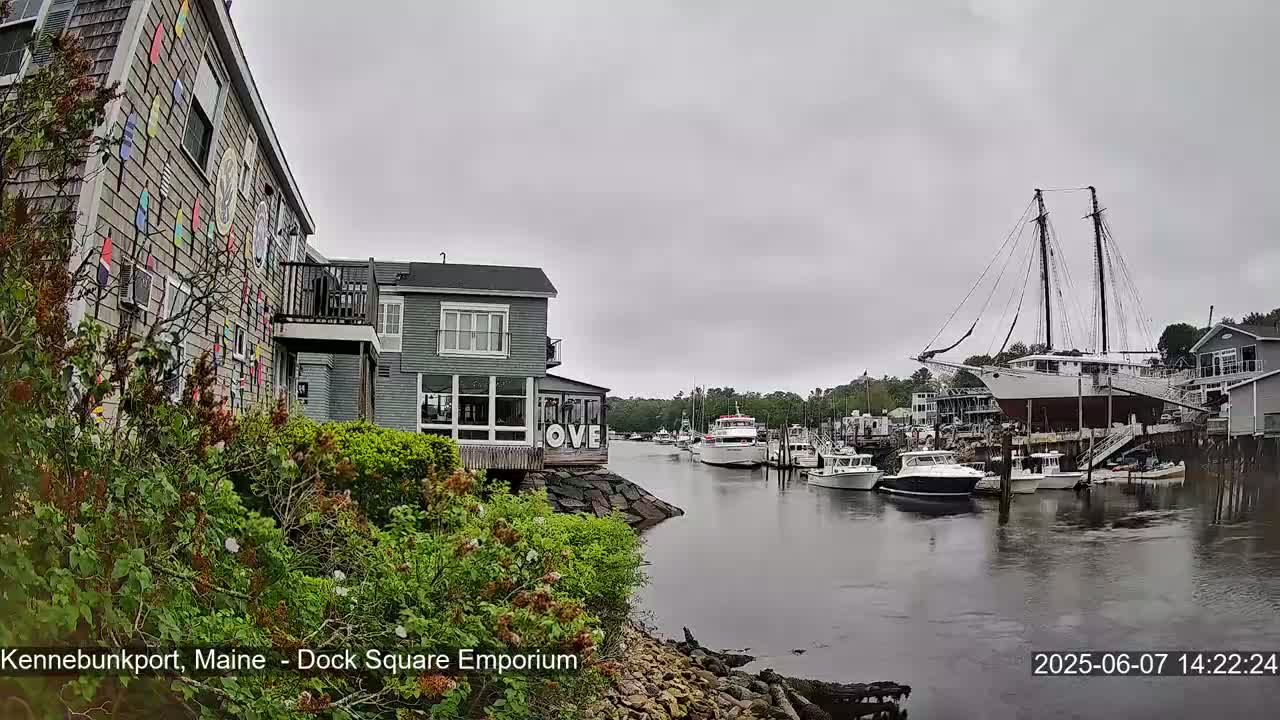 A harbor scene on an overcast day shows several boats, including a large sailboat, moored near buildings with colorful exterior decorations.