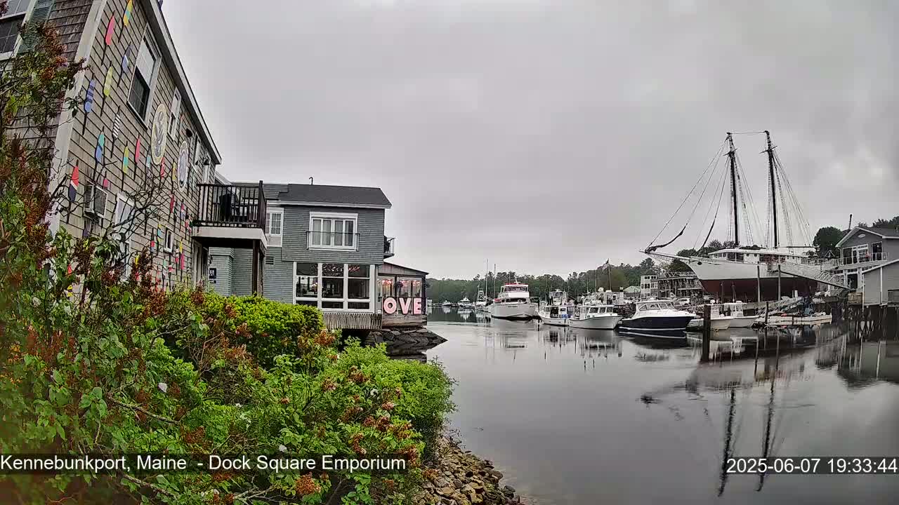 A harbor scene on an overcast day shows several boats, including a large sailing ship, moored along a waterfront with buildings, one of which has colorful nautical flags on its exterior.