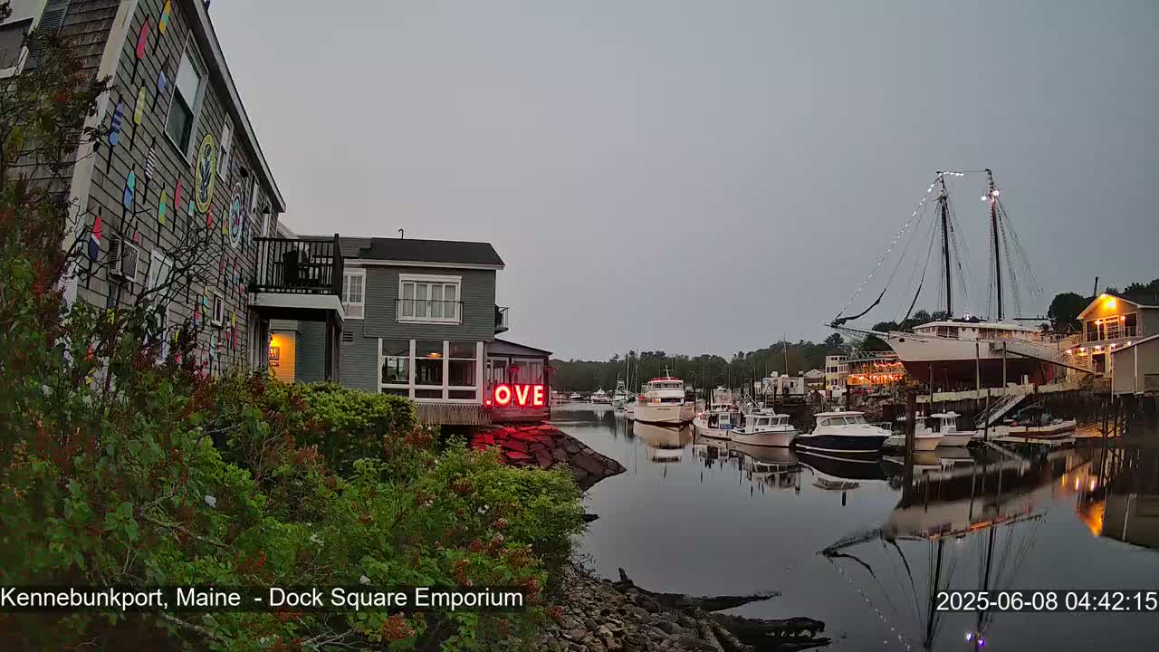 A calm harbor at twilight features a building with a colorful mural, a large sailboat docked, and several smaller boats moored along the water's edge.