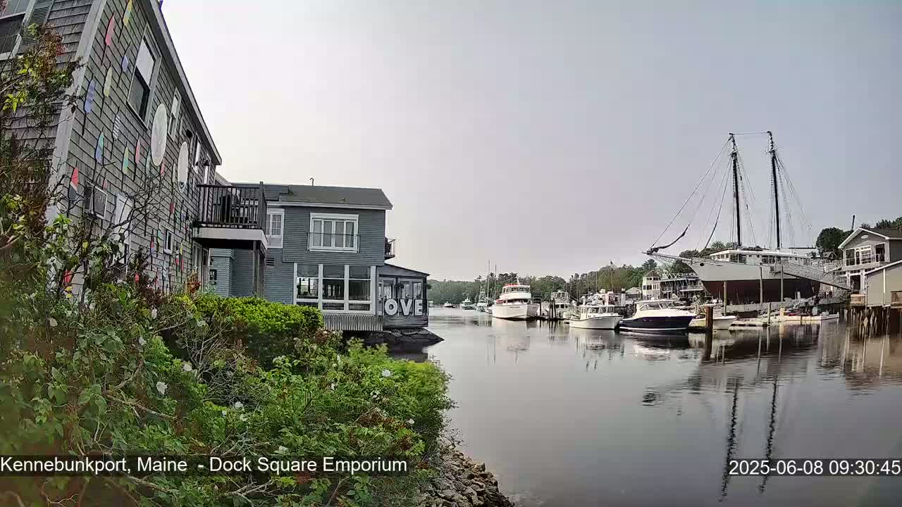 A calm harbor scene shows several boats, including a large sailboat, moored near waterfront buildings under an overcast sky.