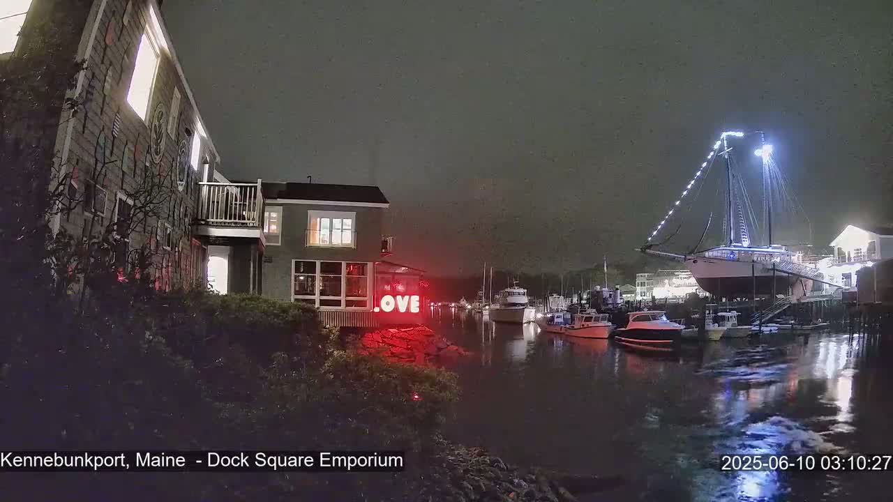 A nighttime harbor scene shows a large building with a red "LOVE" sign, several boats, and a docked sailboat illuminated with white lights, under a dark, cloudy sky.