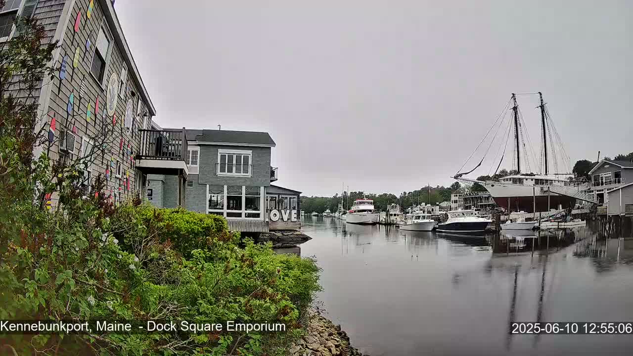 A waterfront scene on an overcast day shows a building decorated with colorful nautical flags next to a calm harbor with several boats and a large sailing ship.
