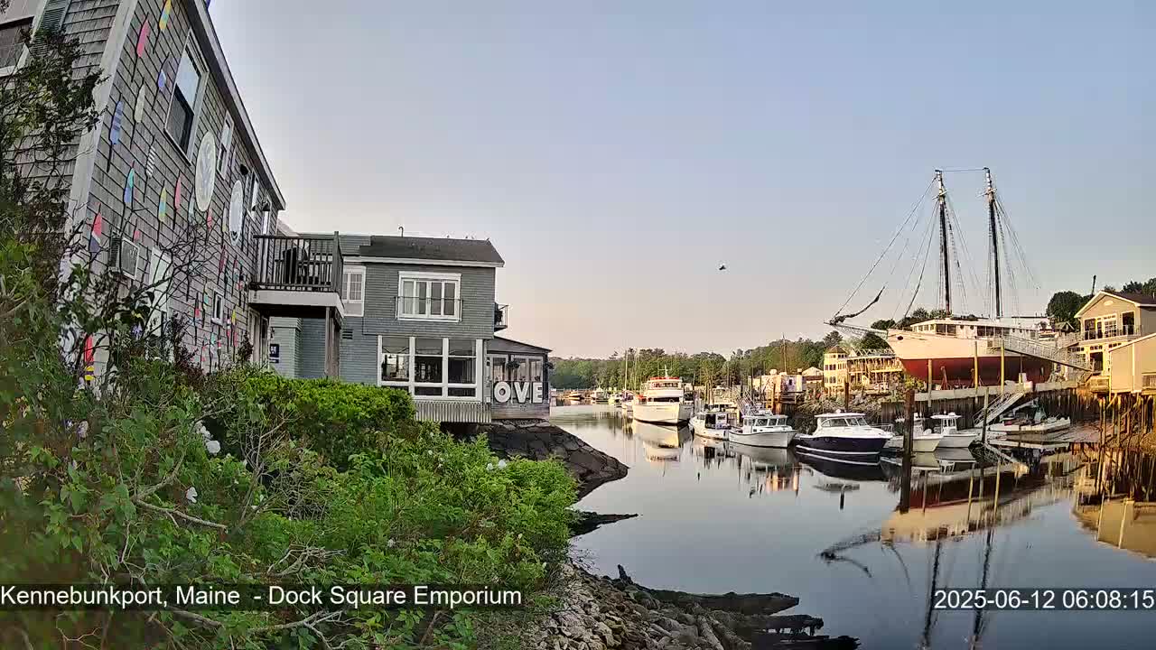 A calm harbor scene under a clear sky features a colorful building, several boats, and a large sailing ship docked near waterfront buildings.