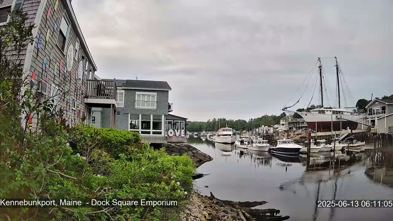 A harbor scene on an overcast day shows several boats, including a large sailing ship, moored along a waterfront with buildings, one of which is adorned with colorful decorations.
